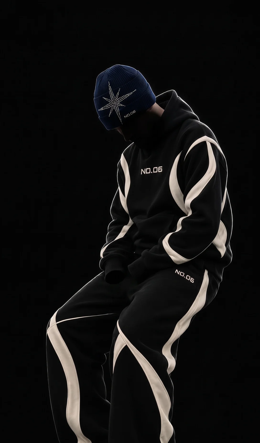 NO.06 Panel Hoodie and Wide Sweatpants in black, seated, on black